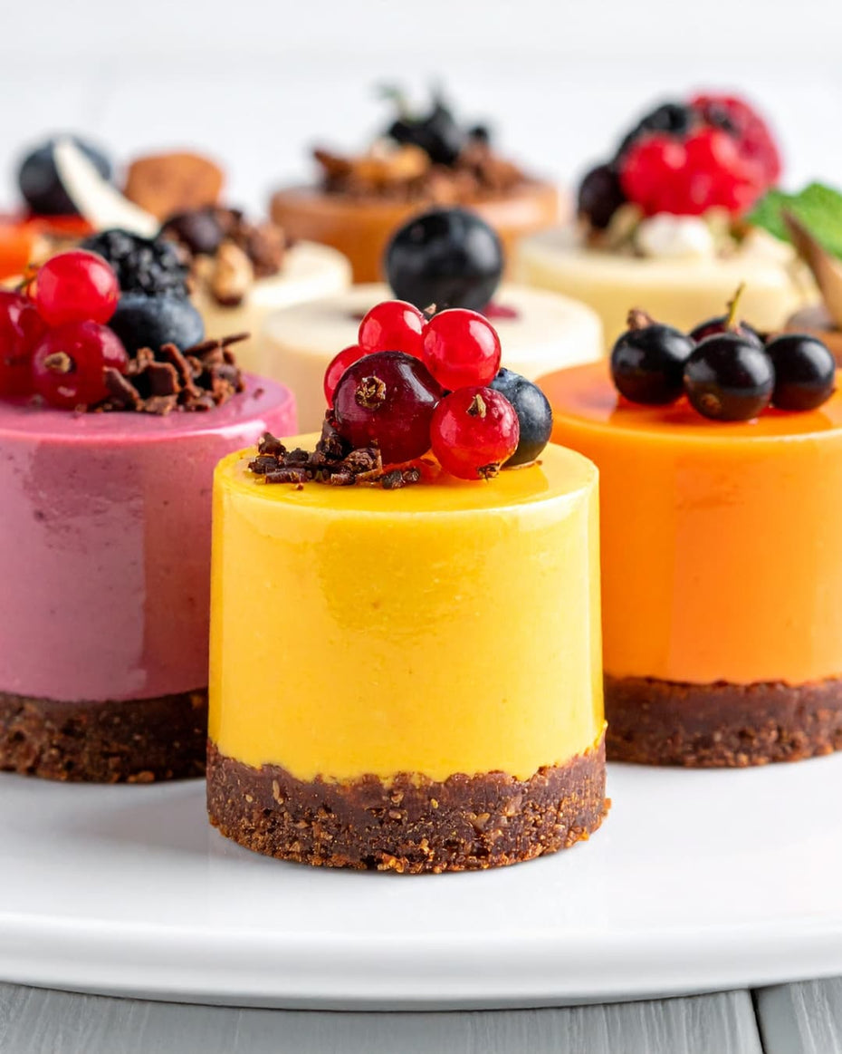 A variety of colorful mini mousse cakes with fresh berry toppings and crushed nuts on individual biscuit bases, part of a gourmet dessert selection for bulk orders.
