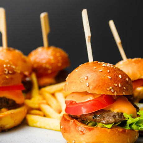 Four mini cheeseburgers with sesame seed buns, wooden skewers, french fries, and tomato slices on a plate against a dark background.

